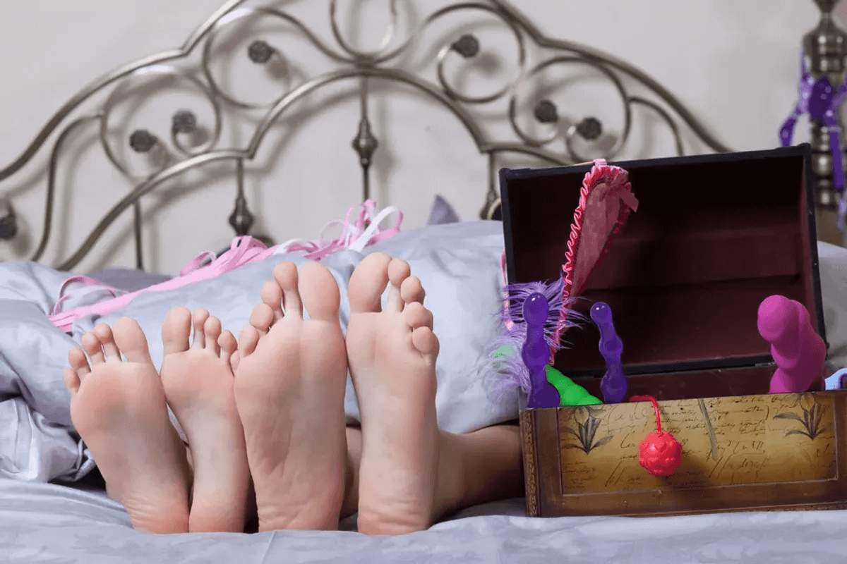 Two pairs of feet from under the sheets with a box of sex toys open besides.