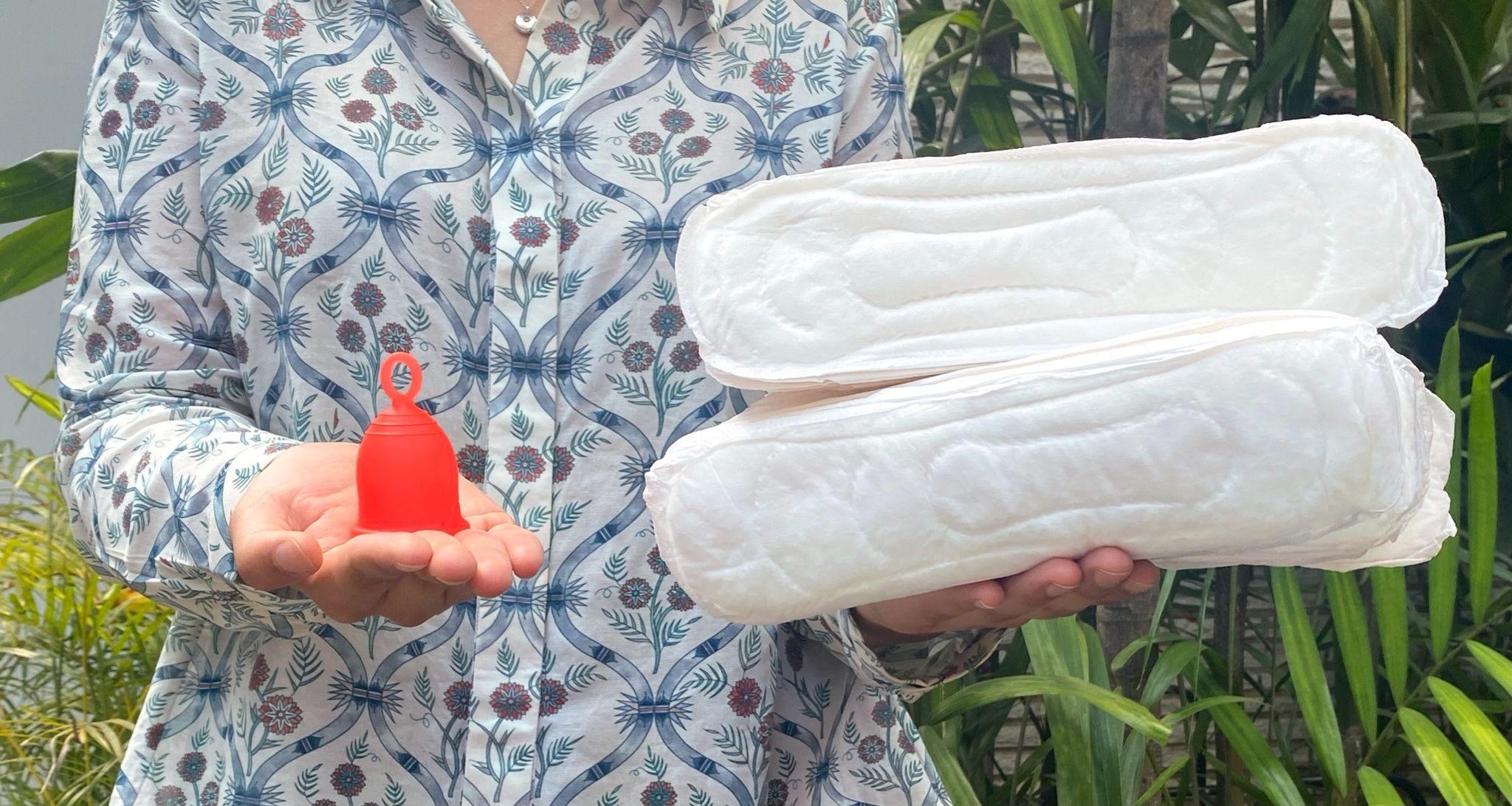Woman holding Asan menstrual cup in one hand and bunch of sanitary pads in other. 